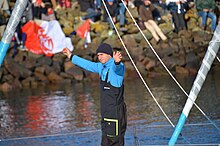 En buste, de trois quarts, debout à l'avant son bateau, les deux bras levés, il salue la foule dans le chenal des Sables-d'Olonne.