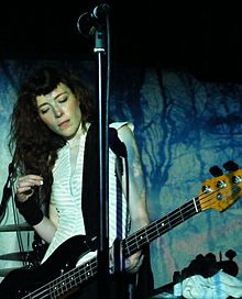 A female musician performing with a bass guitar against a black and blue backdrop. A microphone is visible above her.