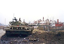 A heavily damaged tank which has lost its right-hand tracks sits next to a mine crater beside a road, with its barrel facing right and ruined houses in the background.