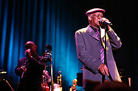 A man wearing a cap on his head, eye glasses, and a suit, holding on to a microphone stand with both hands. To the left of him is a man also holding on to a microphone stand. In between them are various musical instruments.