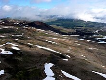 A dark-coloured volcanic cone with a summit crater rising above a sparsely snow-covered plateau with little vegetation.