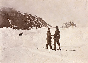 Deux hommes se serrent la main au milieu d'un champ de neige, un chien à leurs côtés. On aperçoit de sombres collines à l'arrière-plan.