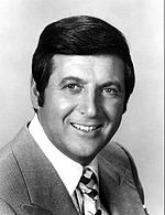 A man with dark hair, wearing a suit with a pattern tie