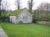 A small, simple chapel seen almost end-on in a grassy burial ground; it is built in stone with a moss-covered roof. On the end is a window with open shutters; on the front face is a door and two shuttered windows.