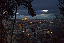 Blick auf die Stadt bei Nacht mit einem angestrahlten Dom. In einzelnen Häusern ist Licht zu sehen und ein heller Mondschein dringt durch die Wolken hervor. Im Vordergrund sind die Silhouetten von Bäumen zu erkennen.