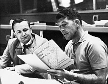 Two men seated at a desk, both reading from a large book the younger man is holding