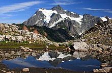 A large and sharp mountain, partly covered in snow, is reflected in a tarn in the foreground.