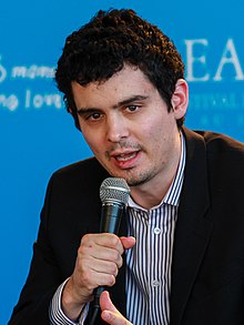 Damien Chazelle de face, la bouche ouverte et un micro en main, habillé d'une chemise et d'une jaquette et devant un fond bleu.