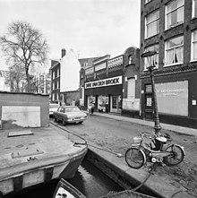 Cliché en noir et blanc d'un alignement de bâtiments, où domine la brique, le long d'un canal : l'un d'eux est une ancienne salle de cinéma de style mauresque où est aménagé un supermarché à l'enseigne Dirk van den Broek.