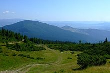 Farbfotografie mit Blick von einer Berghöhe hinunter ins Tal und über weitere Bergketten als Silhouetten im Hintergrund. Der Himmel ist sonnig und hellblau. Eine Alm im Vordergrund auf dem ein Pfad hinunter zu Bäumen und einer Hütte in der Bildmitte führt.