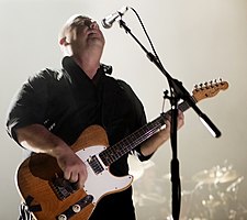 Black Francis playing guitar onstage in front of a microphone, looking up