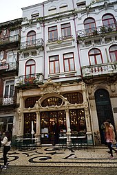 Façade d'immeuble blanc ancien avec des balcons. Au rez-de-chaussée, la devanture stylisée et ouvragée de l'établissement, en pierre de couleur crème avec des fenêtres marron. Il y a deux statues en hauteur et des colonnes de pierre de chaque côté de l'entrée.