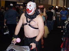 A shirtless man wearing a black leather harness and clown makeup. He has nipple piercings and a tattoo on his chest that is obscured by the harness.