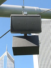 A round, grey metal tube with two dark grey rectangular boxy speakers hanging from it. In the background are the tops of two skyscrapers.