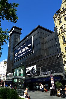 Entrée d'une salle de cinéma dans un édifice noir surmonté d'une tour à gauche.