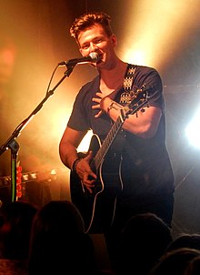 Tyler Ward wearing dark clothing, standing onstage with an acoustic guitar, appearing to sing or speak into microphone