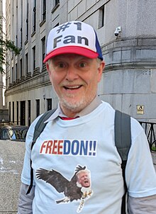 Barbieri as his character Jake Byrd at the Trump criminal trial in Manhattan. Byrd's eyebrows are raised unevenly while obscured by the brim of his cap with an almost surprised childish grin. He has a white and grey close cropped mustache and beard that is somewhat patchy. Byrd is wearing a red, white, and blue baseball cap that reads "#1 Fan" in black block letters against a white background. Byrd is also wearing a backpack over a light blue colored t-shirt that Byrd is wearing over tan colored collared shirt with a red necktie (only an accent of the red tie is visible) the shoulder straps of the backpack frame his necktie and chest. On the light blue (outermost) t-shirt is the word: FREEDON!! (a play on FREEDOM) the text is on a white background with the FREE in white and the DON!! in blue. Beneath the FREEDON!! text is an image of a bald eagle with Donald Trump's head superimposed over the eagle's head. Trump's expression is ambiguous. He is squinting and his mouth is wide open; he could be yelling in pain or attempting to incite a mob. The eagle's right talon is handcuffed, but the chain of the handcuff is broken at the seventh link.