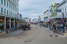 A street, surrounded on both sides by two- and three-story buildings. One person is riding on horseback in the middle of the street, while others are walking on the sidewalk. Bikes are parked at the curb.