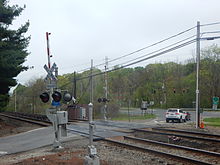 Train tracks going from lower right to mid-left cross, at grade, with metal plating around the tracks, a paved road going lower left to an intersection at a divided highway with a traffic light just above the tracks. Two railroad crossing signs with red-and-white-striped barriers in the up position are on either side of the tracks. A pine tree's branches protrude into the image from the left; across the divided highway are more woods, with trees beginning show leaf, slightly mited under an overcast sky