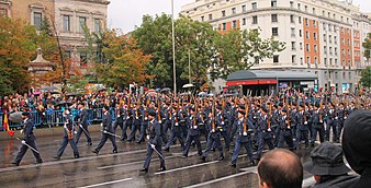 Varias docenas de soldados masculinos en uniformes formales de color azul acero, portando rifles de madera, marchan por una amplia calle mientras una multitud observa