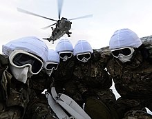 Colour photograph of five fully covered marines in a huddle underneath a helicopter.