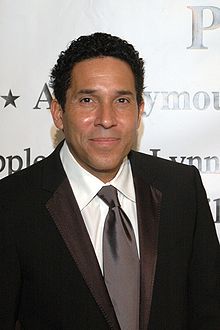 Close-up of Óscar Núñez wearing a black suit and tie with white shirt, smiling at camera