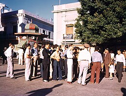 Une place ensoleillée avec des personnes de type latino vêtues plutôt élégamment dans le style années 30.