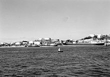 Black and white photograph of three large motor ships docked next to an island covered in large white storage tanks. A small boat is visible in the foreground.