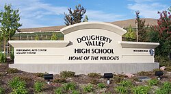 A beige school sign, reading "Dougherty Valley High School", and then underneath in smaller italicized text, "Home of the Wildcats". To the left are even smaller texts reading "Performing Arts Center" and "Aquatics Center", and to the right are the school's address and a wildcat logo.