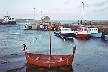 A photograph of a red boat floating in water, with several boats in a pier in the background