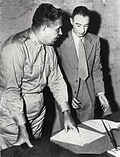 A man in shirt and tie and another wearing a suit stand behind a writing desk. On the wall behind is a map of the Pacific.