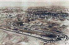 Image en noir et blanc. Au fond, la ville de Los Angeles. À l'avant-plan, un immense ovale bordé de petites maisons de plain-pied, qui forment le village olympique ; l'intérieur de l'ovale contient trois bâtiments en longueur et de la verdure.