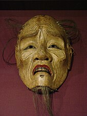 Masque en bois non peint, sauf les lèvres rouges, d'un vieillard émacié et ridé avec une barbiche et de rares cheveux.