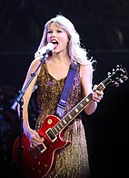 Swift playing a red electric guitar while singing onto a mic