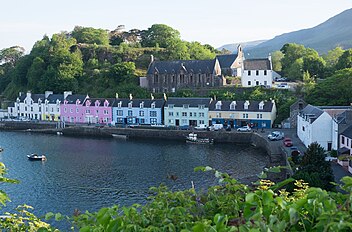 A small harbour fronted with a row of cottages painted in white, pink, green and blue with a tree-covered hillock behind them.