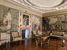 A photograph of a richly-decorated room, with walls covered by tapestries and several pieces of sitting room furniture as well as an elaborately-carved ceiling