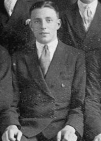 A man with dark hair in a suit and tie sits with his hands on his knees