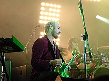 Johns holds his plectrum away from the guitar. He is bearded and shown in right profile. Gillies is partly obscured by his drum kit. Joannou is not in view.