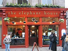 Façade d'un pub intégralement rouge avec une porte d'entrée centrale située en renfoncement. On aperçoit des gens attablés à travers la vitre de gauche et le nom « the elephant house » est écrit en lettres dorées en haut de la façade, sous des jardinières fleuries. Sur la droite, des personnes s'attardent à l'extérieur en observant la vitrine