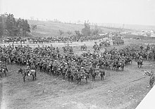 A large group of men and horses drawn up into lines in a field and on the adjoining road. A hill with trees and tents can be seen in the background.