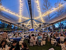 Pomona alumni eating on Marston Quad at round blue tables with plastic chairs under a translucent tarp with string lights