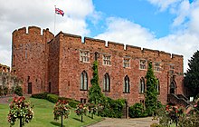 Red stone built castle with turret on the left and flowers lining the approach road