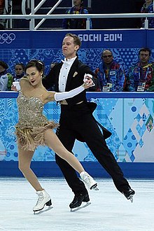 Chock und Bates auf der Eisfläche während ihres Kurztanzes. Bates trägt einen schwarzen Anzug, darunter ein weißes Hemd mit goldenen Details; Chock trägt ein an ein Abendkleid angelehntes goldenes Kleid, lange weiße Handschuhe und goldene Armreifen. Im Hintergrund ist die Aufschrift "Sochi 2014" zu sehen.