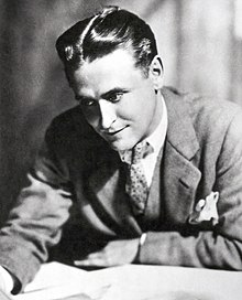 A photograph of F. Scott Fitzgerald by Nickolas Muray. Fitzgerald is bent over a desk and is examining a sheaf of papers. He is wearing a light suit and a polka-dot tie. A white handkerchief is in his breast pocket.