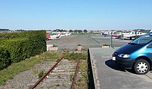 Vue de la voie désaffectée traversant le tarmac de l'aéroport, avec sur la droite l'ancien quai de la halte (délimitant un parking).