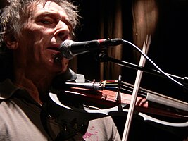 Cale playing a viola while singing into a microphone onstage