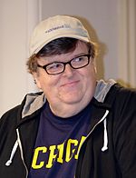 A man standing in front of a white wall is wearing a beige baseball cap over his head. He is also dressed in a black T-shirt with yellow lettering underneath a black hooded jacket with white drawstrings.