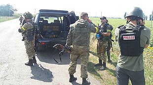 Uniformed soldiers, a journalist and a vehicle at the side of a road