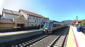 A train at a high-level platform, with a mission-style depot building behind