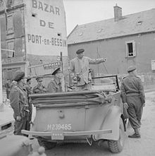 Photographie montrant un véhicule décapoté occupé par deux personnalités dont une debout et entouré de cinq personnes.